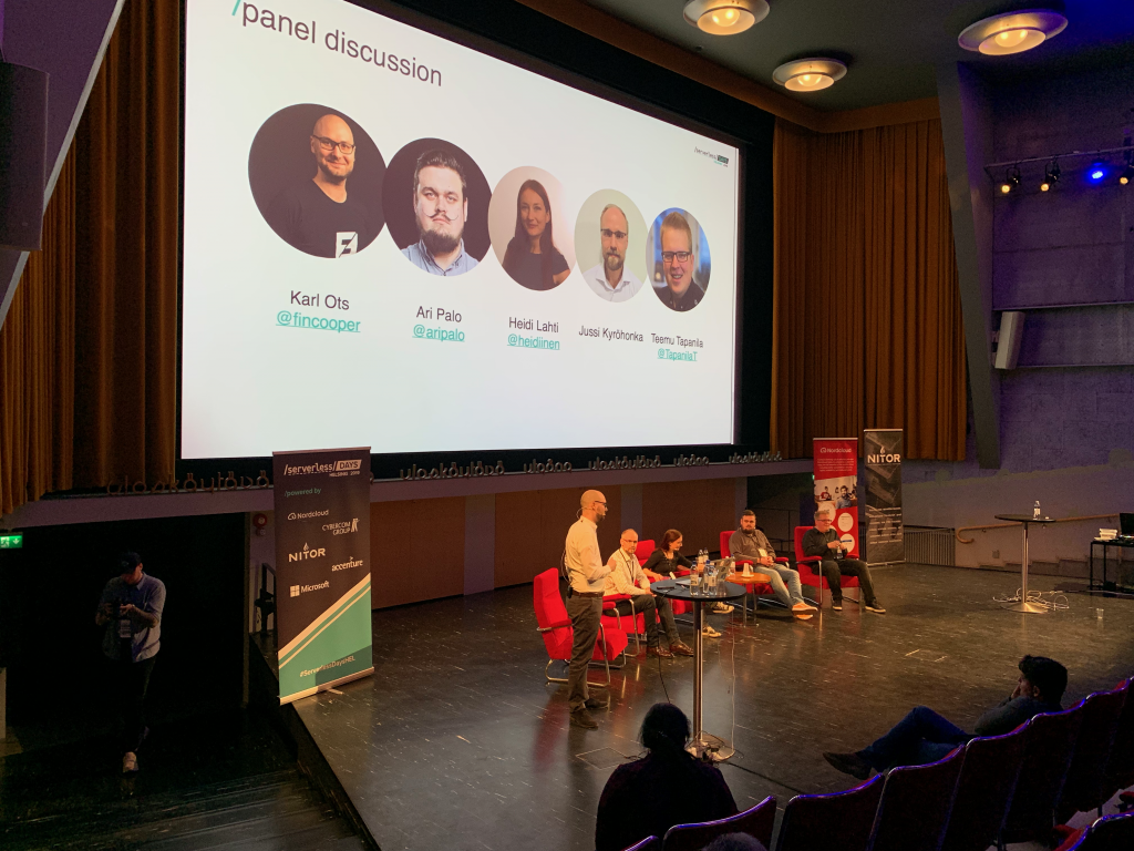 Panelists (Jussi, Heidi, Ari and Teemu) sitting at the stage with moderator Karl standing on the left